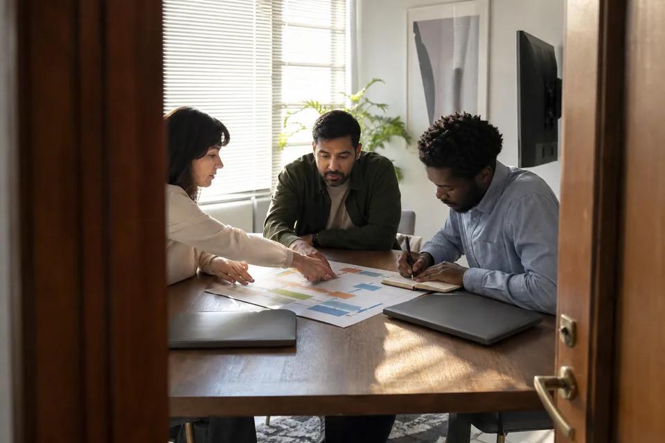 A small marketing team at a table in daylight, two people pointing at a printed plan while a third takes notes, monitors turned away