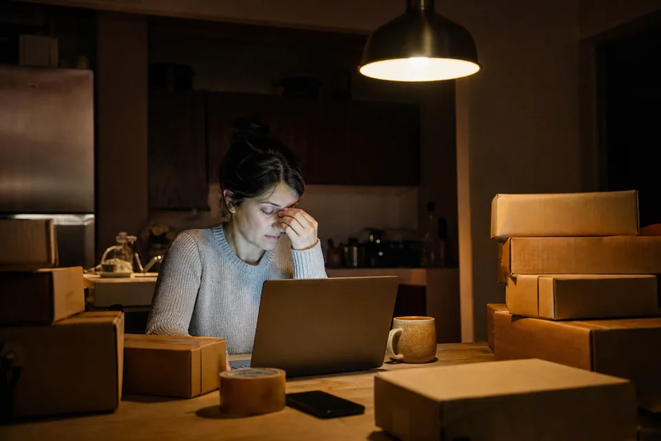 A shop owner at a kitchen table late in the evening with a laptop, parcels and a cold cup of coffee, one lamp on